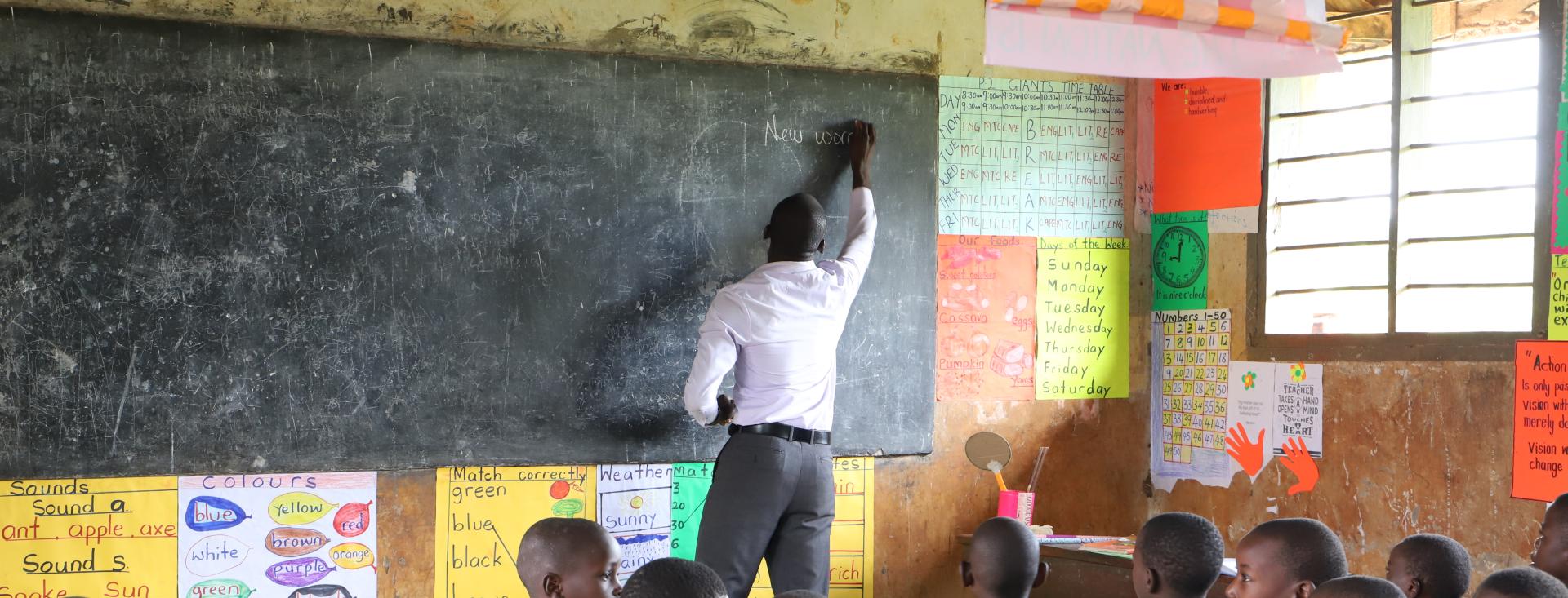 A young Black man in a white shirt writes on a chalk board with his back to a class of children seated in desks