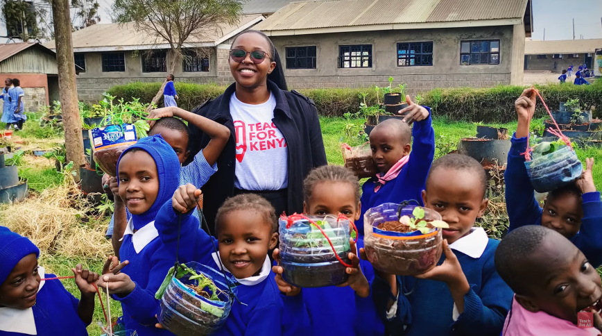 A Black female teachers stands in a school yard surrounded by young students holding up plants growing in recycled plastic containers