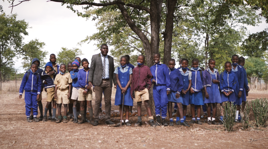 In front of a large tree, a tall African man stands in the middle of a row of African students in school uniforms